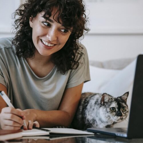 Glad woman with cat writing in planner while using laptop
